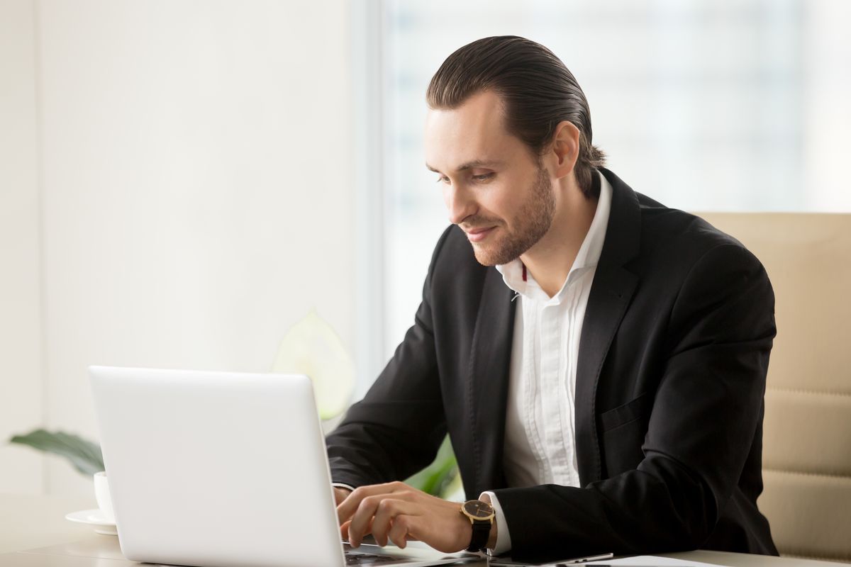 A practice owner in a blazer smiling while typing on a laptop at a bright office desk