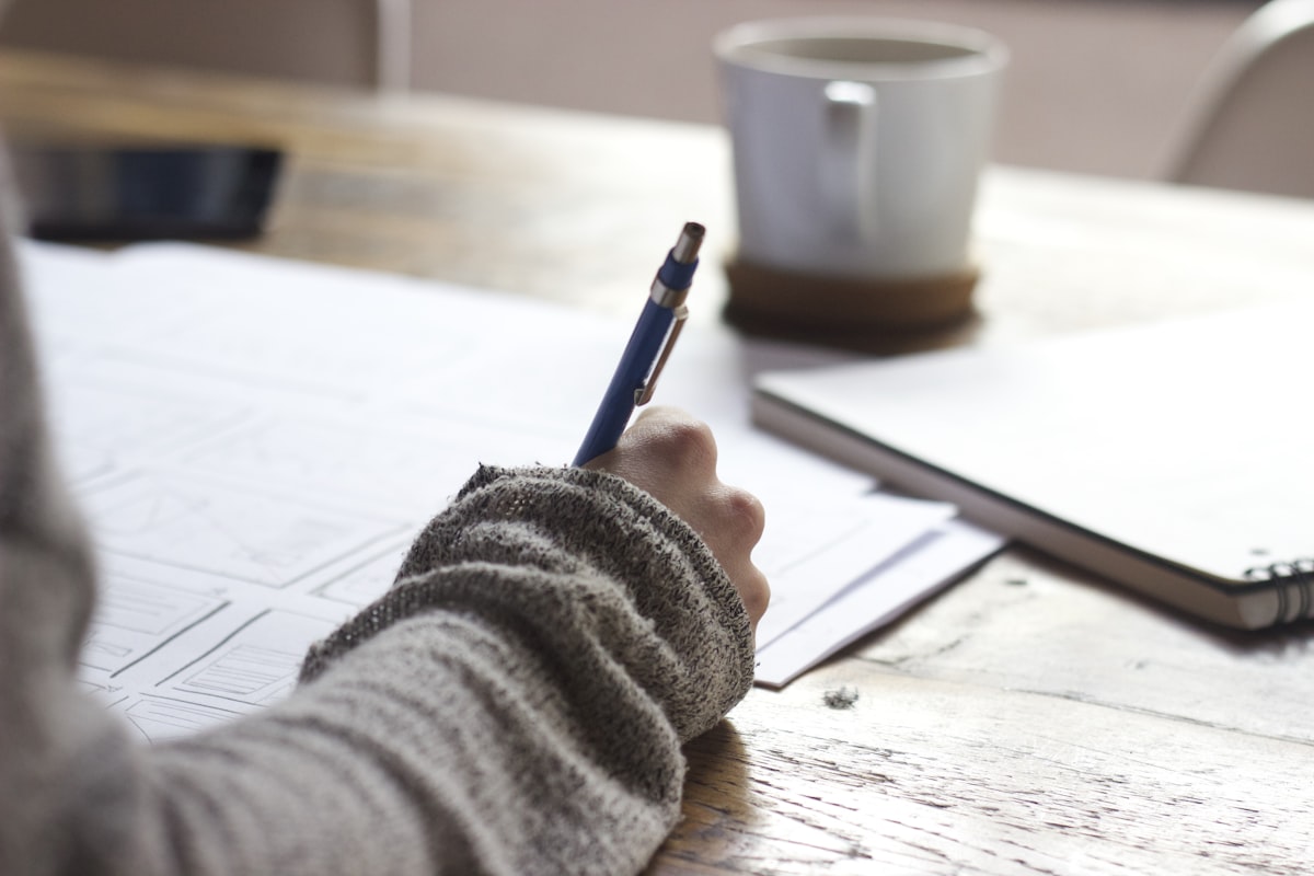 Person working through printed pages with a pen beside a coffee cup and notebook