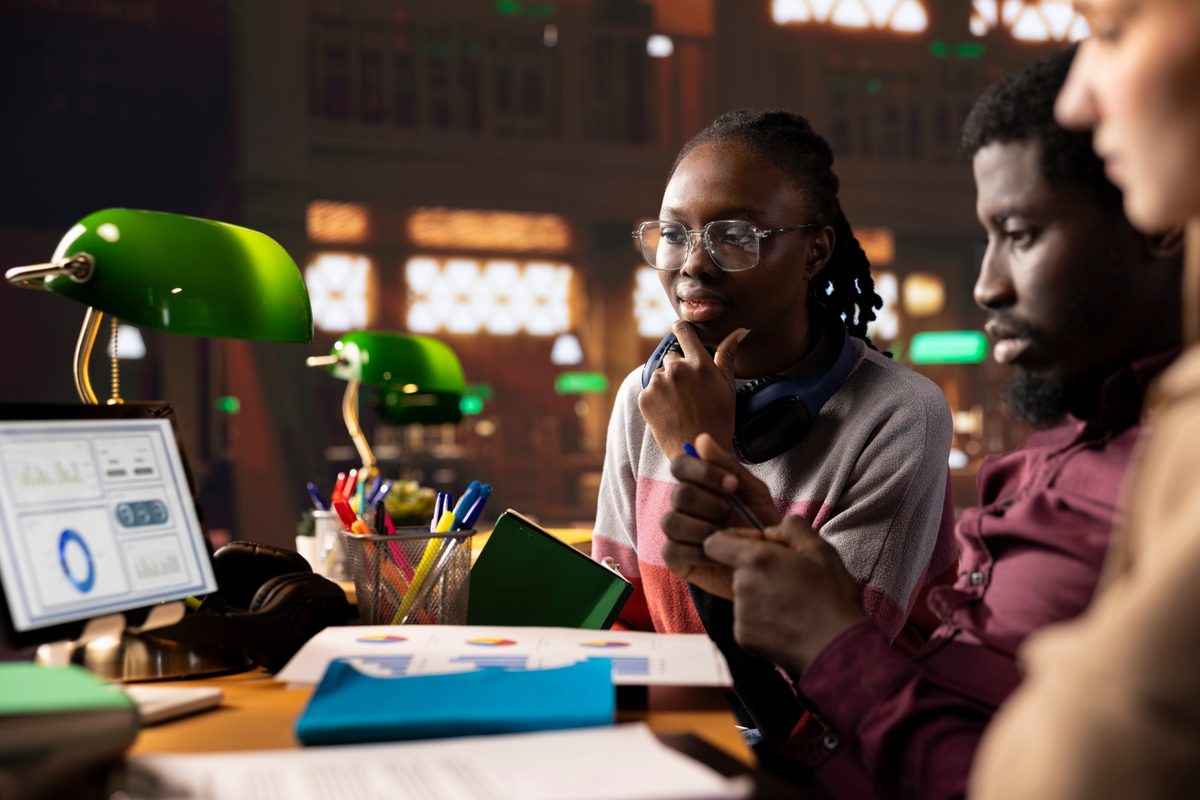 Three founders at a desk under green banker's lamps reviewing printed charts and a dashboard on a monitor